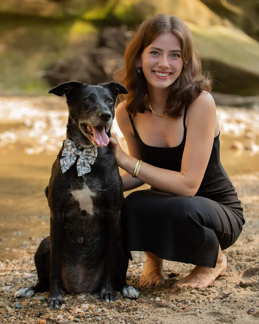Brookelyn crouching beside a black dog in a bow tie on a sandy creek bank