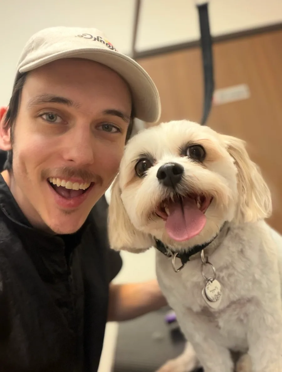 Logan smiling beside a small cream-colored dog he just groomed